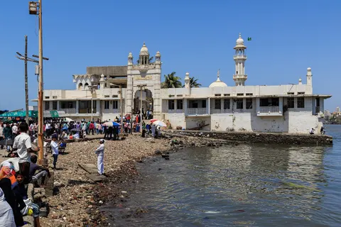 The dargah’s white gateway and minaret beside the water, with visitors on the rocky shore